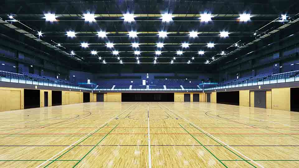 A large, empty indoor sports arena featuring a polished hardwood floor with multi-colored court lines for various sports